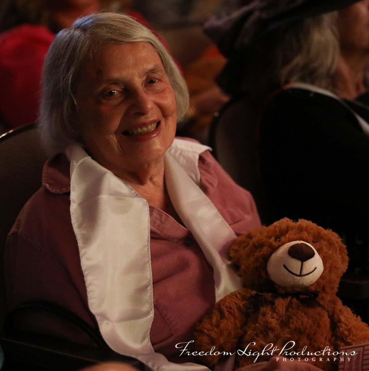 Smiling elderly woman with gray hair sitting in a dimly lit theater, holding a brown teddy bear, wearing a pink shirt with a white scarf, and sitting next to a person with curly hair and a hat.