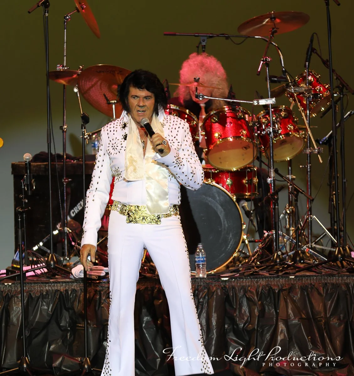Elvis Presley performing on stage with a microphone in his right hand, wearing a white jumpsuit with gold and black embellishments, and a drummer playing behind him with a red drum set.