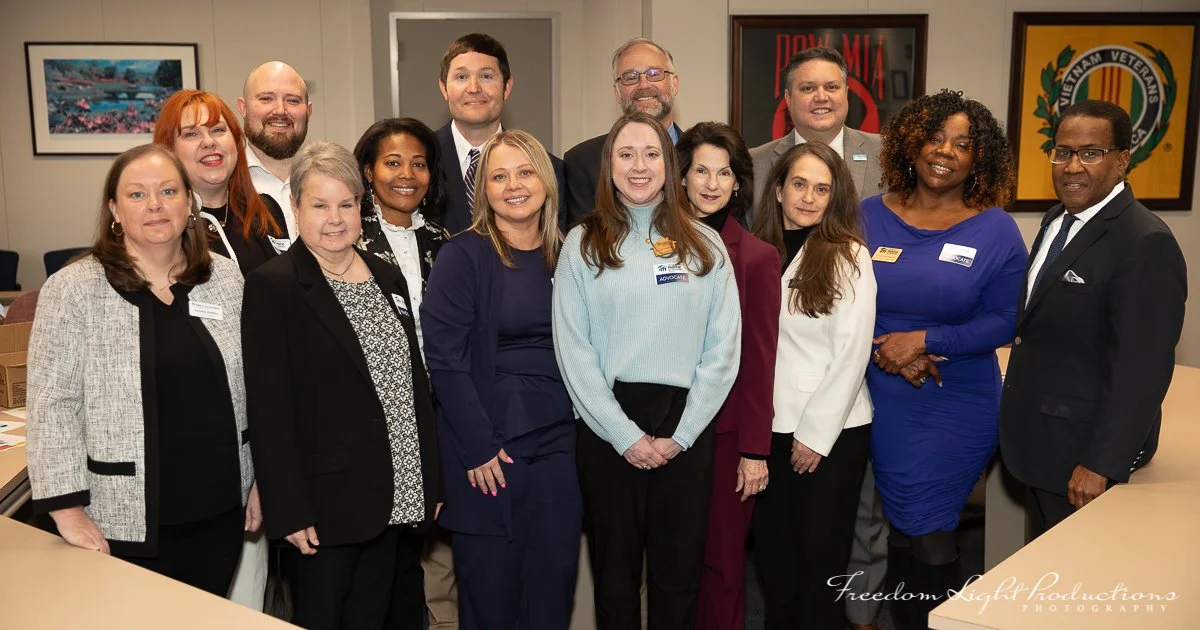 A group of fifteen people, mostly women, standing together in an indoor office or conference room, smiling at the camera. There are framed pictures and posters on the walls behind them.