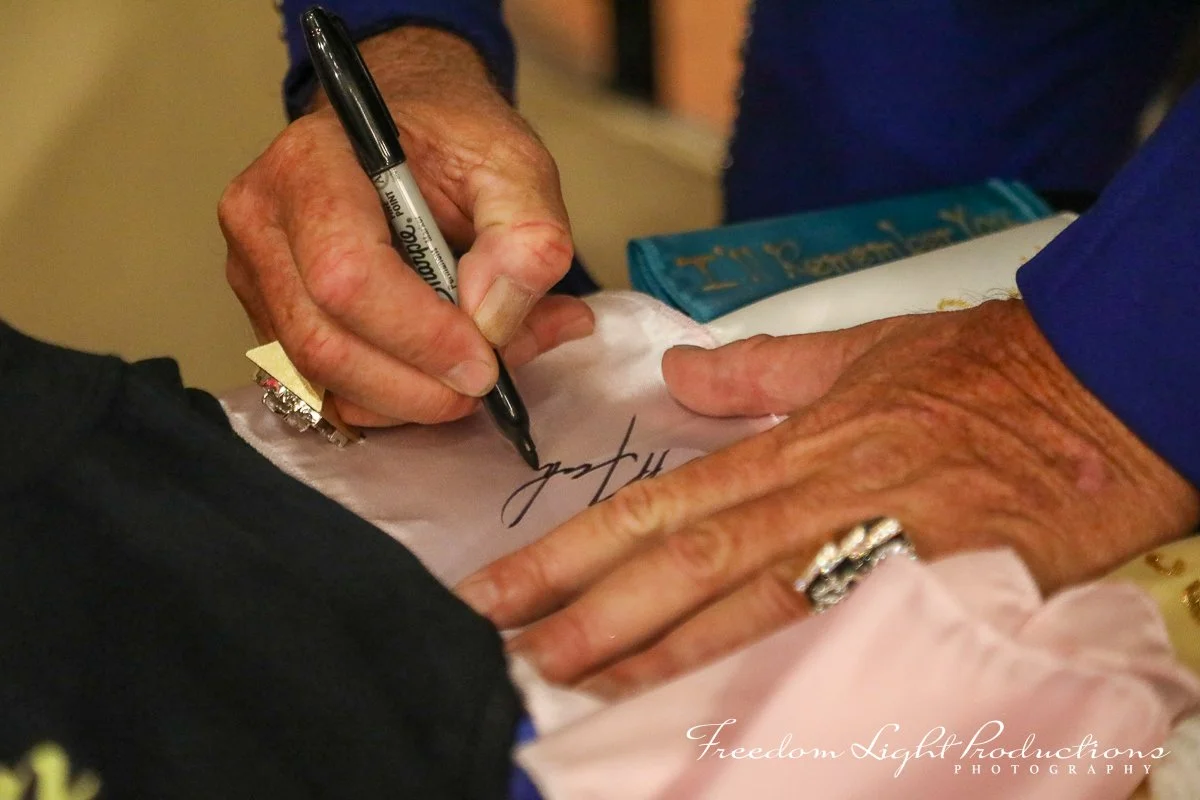Close-up of an elderly person signing a paper with a black marker, with another hand resting on the paper, wearing a ring, and a blue cloth visible in the background.