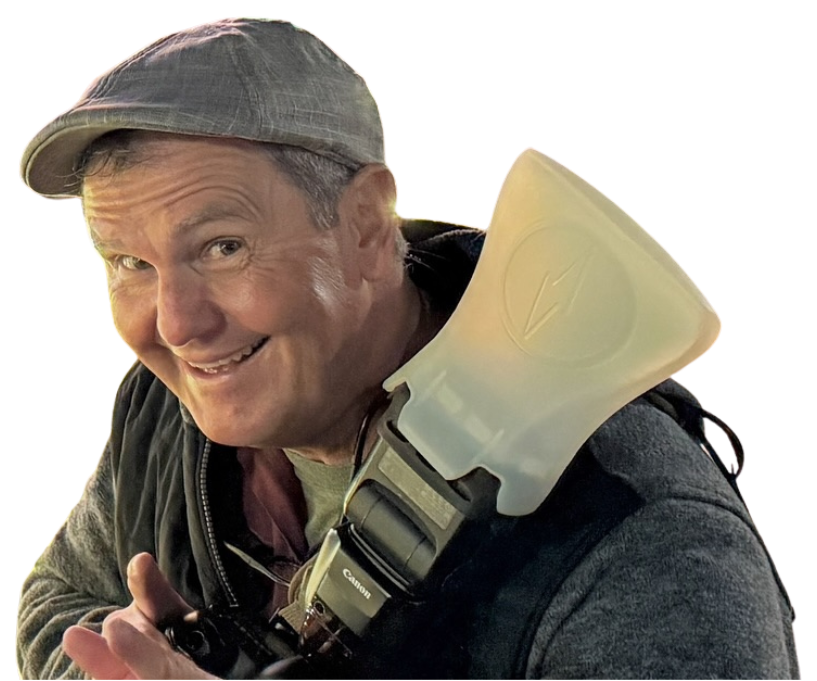 A smiling man wearing a gray cap and a dark jacket, holding a camera with a large flash attached, against a colorful background.