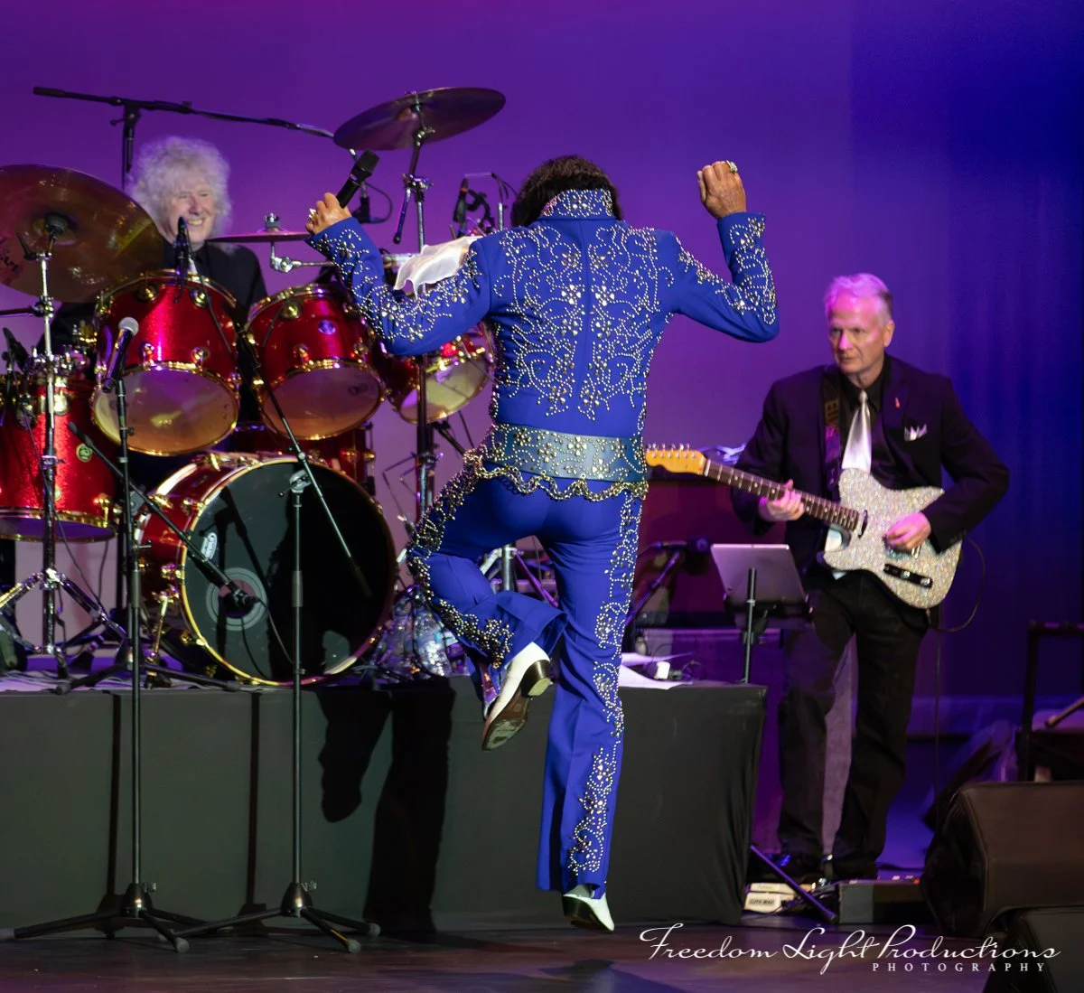 Performance on stage with a drummer, a guitarist, and a singer dressed in a blue jumpsuit with intricate white and gold embellishments. The singer is mid-dance with one leg raised, and the guitarist is playing guitar. The scene is colorful with stage