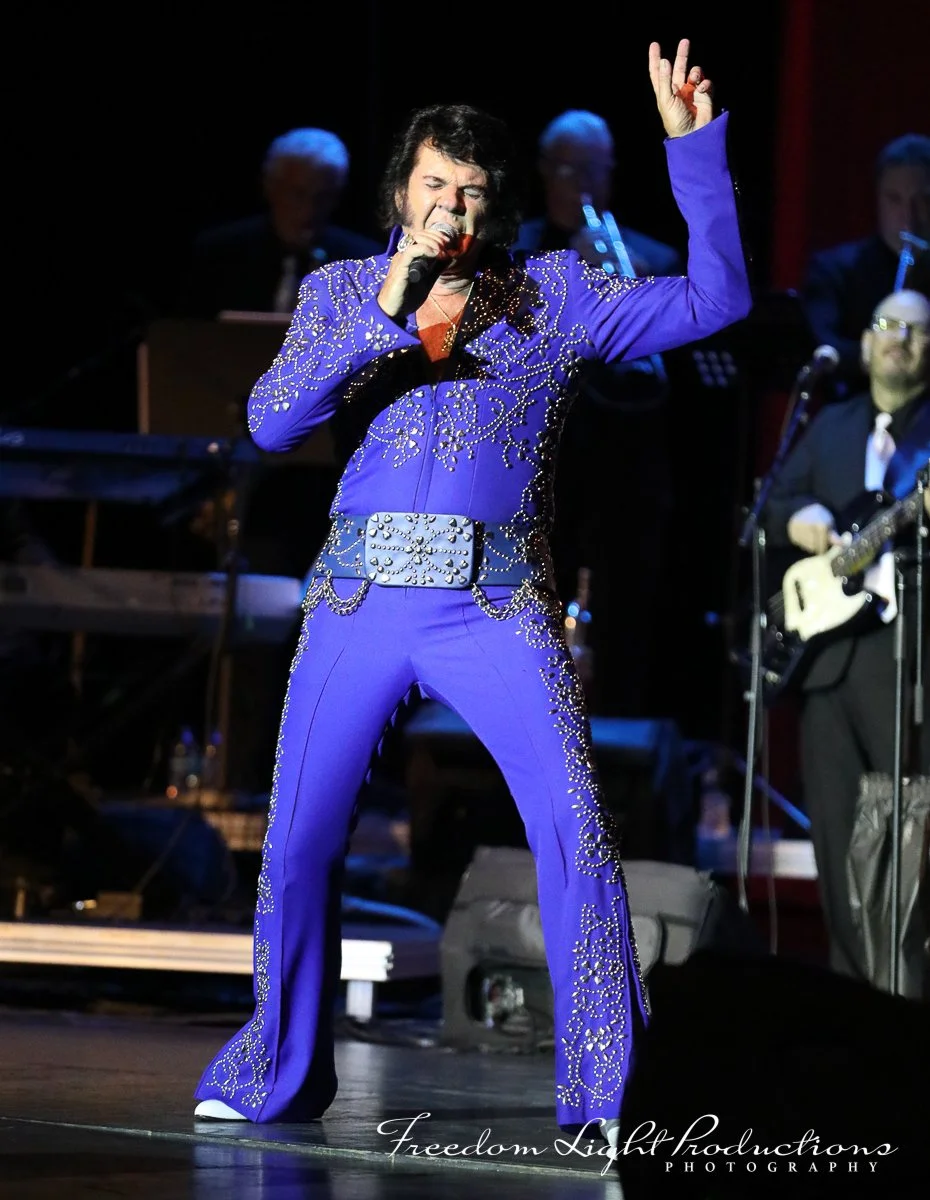 Performing Elvis Presley in a blue, rhinestone-studded jumpsuit with a large belt, singing into a microphone, with musicians playing instruments in the background.