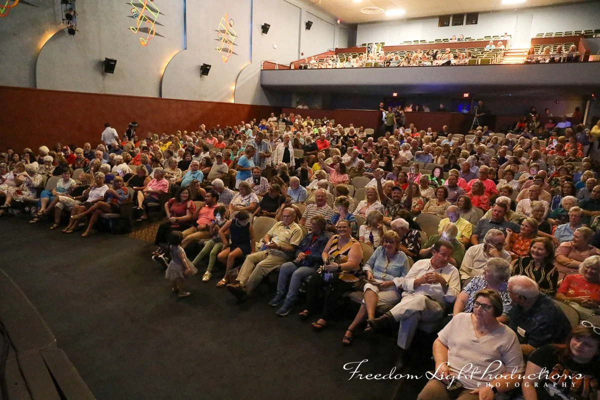 Audience seated in a theater or auditorium, some smiling and engaging with an event on stage, with a few children walking in front.