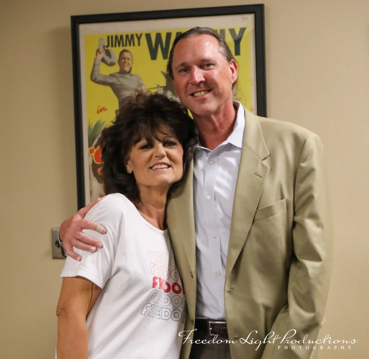 A woman and a man posing together in front of a vintage Jimmy Wagy poster. They are smiling and embracing each other.