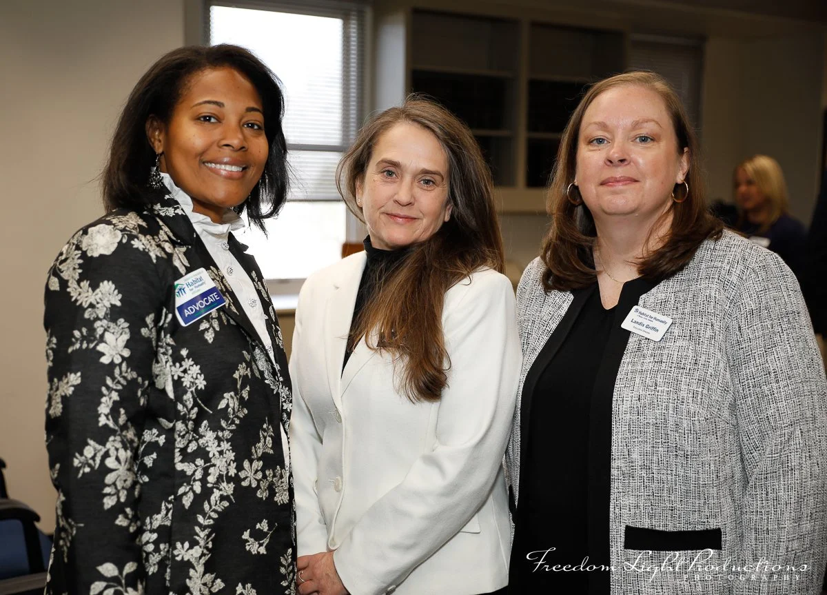Three women standing together in an indoor setting, smiling at the camera. The woman on the left wears a floral blazer with an 'Advocate' badge, the woman in the middle wears a white blazer, and the woman on the right wears a black top and a gray bla