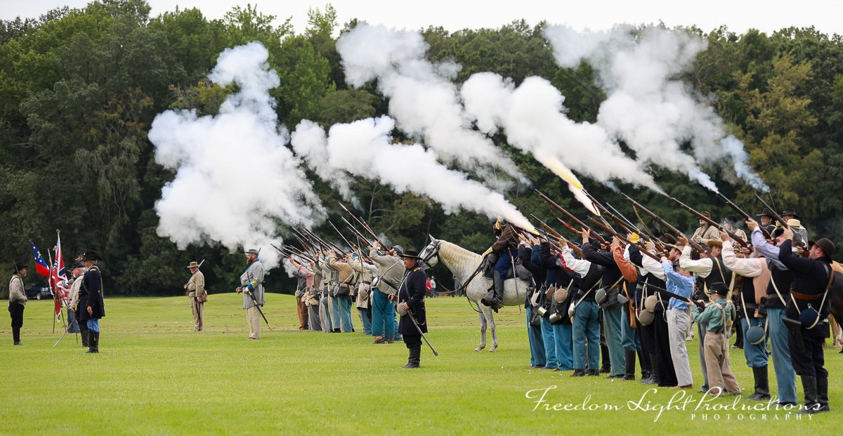 Civil War reenactment with soldiers in period uniforms firing muskets in a field, with flags and smoke visible.