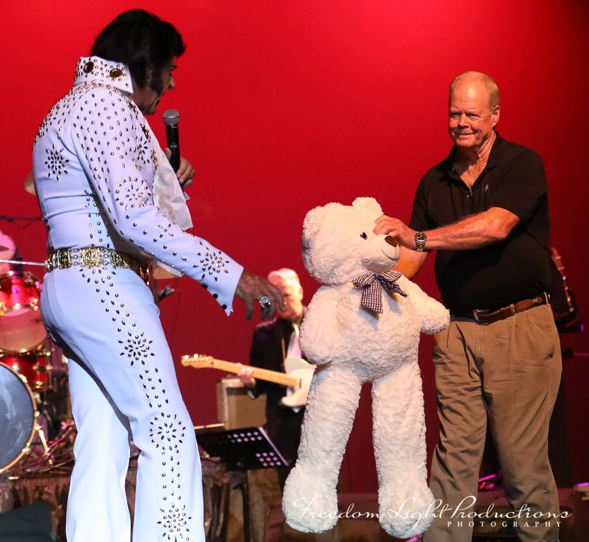 Elvis Presley impersonator in a white, studded jumpsuit holding a microphone, and a man in a black shirt and khakis holding a teddy bear on stage with a red backdrop.