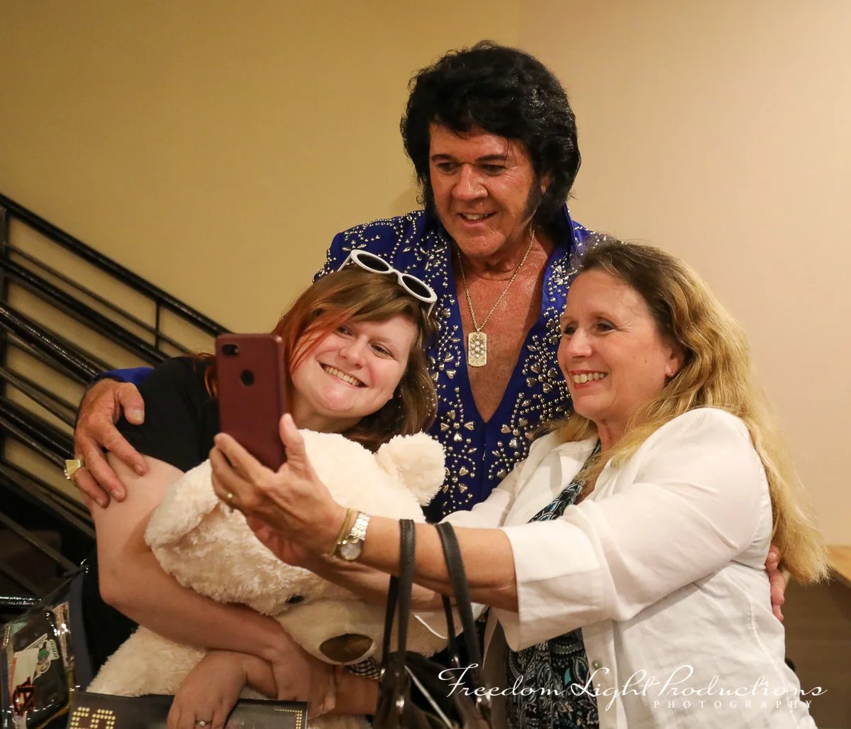 Three people taking a selfie together: a woman on the left holding a teddy bear, a man in the middle wearing a bright blue shirt with rhinestones, and a woman on the right in a white blazer. They are smiling and posing for the photo indoors.