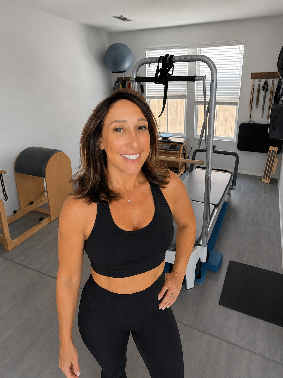 A woman in workout clothes smiling in a home gym, with fitness equipment like a pilates reformer, exercise ball, wall-mounted hooks with workout accessories, and large windows with blinds.