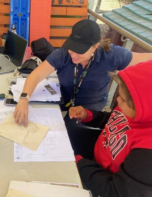 A woman in a black cap and blue shirt and a student in a red hoodie are examining design plans on a table at an outdoor setting.