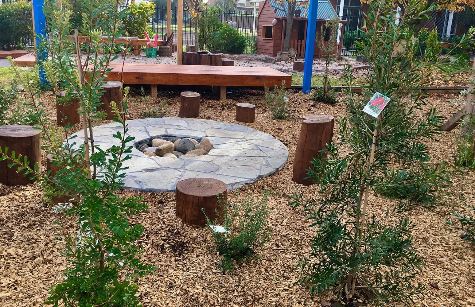 A children's outdoor playground with a circular stone fire pit surrounded by wooden stumps for seating. There are plants and small trees with labels, a wooden raised platform, and a playhouse in the background.