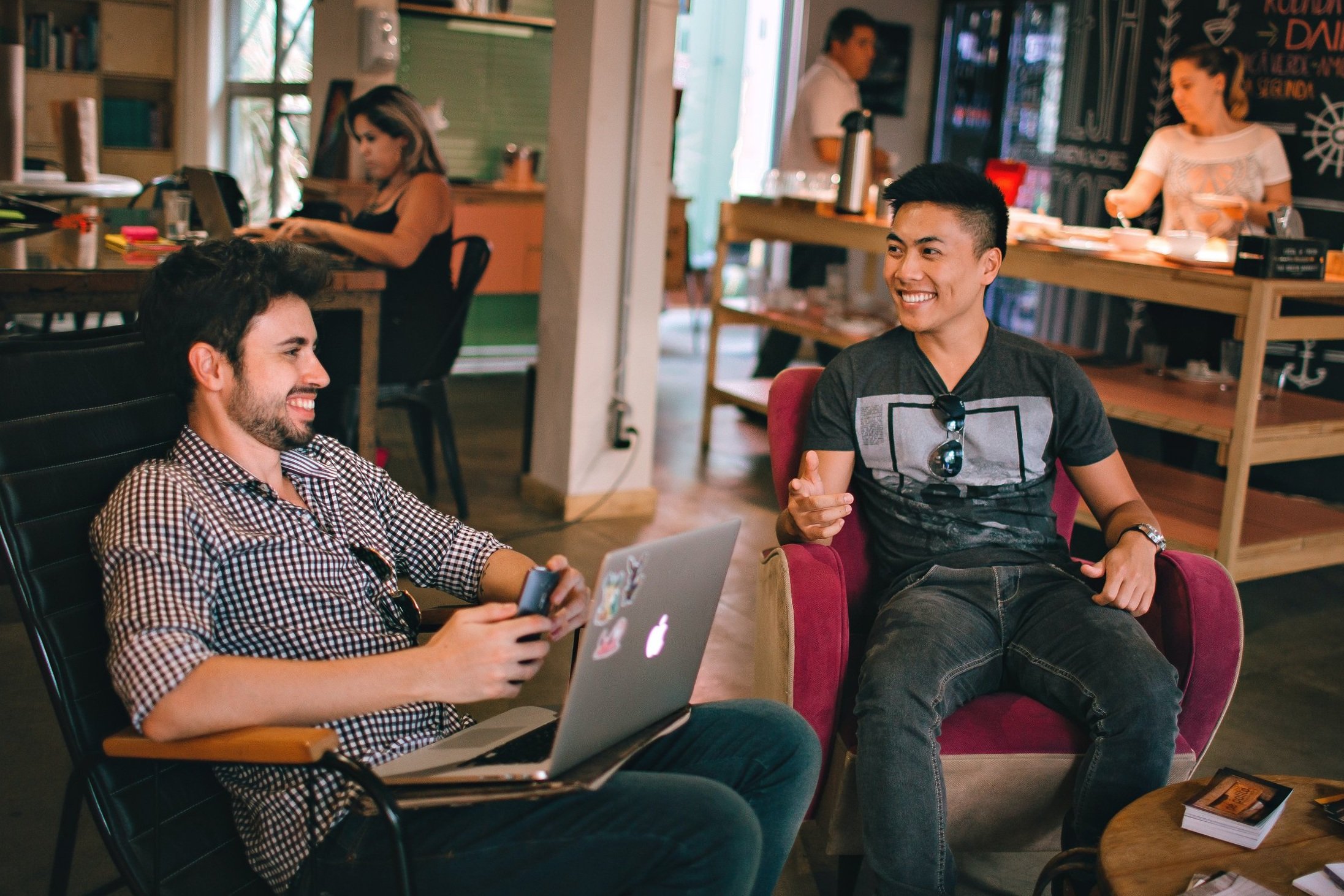 Two people sitting and smiling in a cozy cafe, one using a laptop, the other holding a cup. Two more people working in the background.