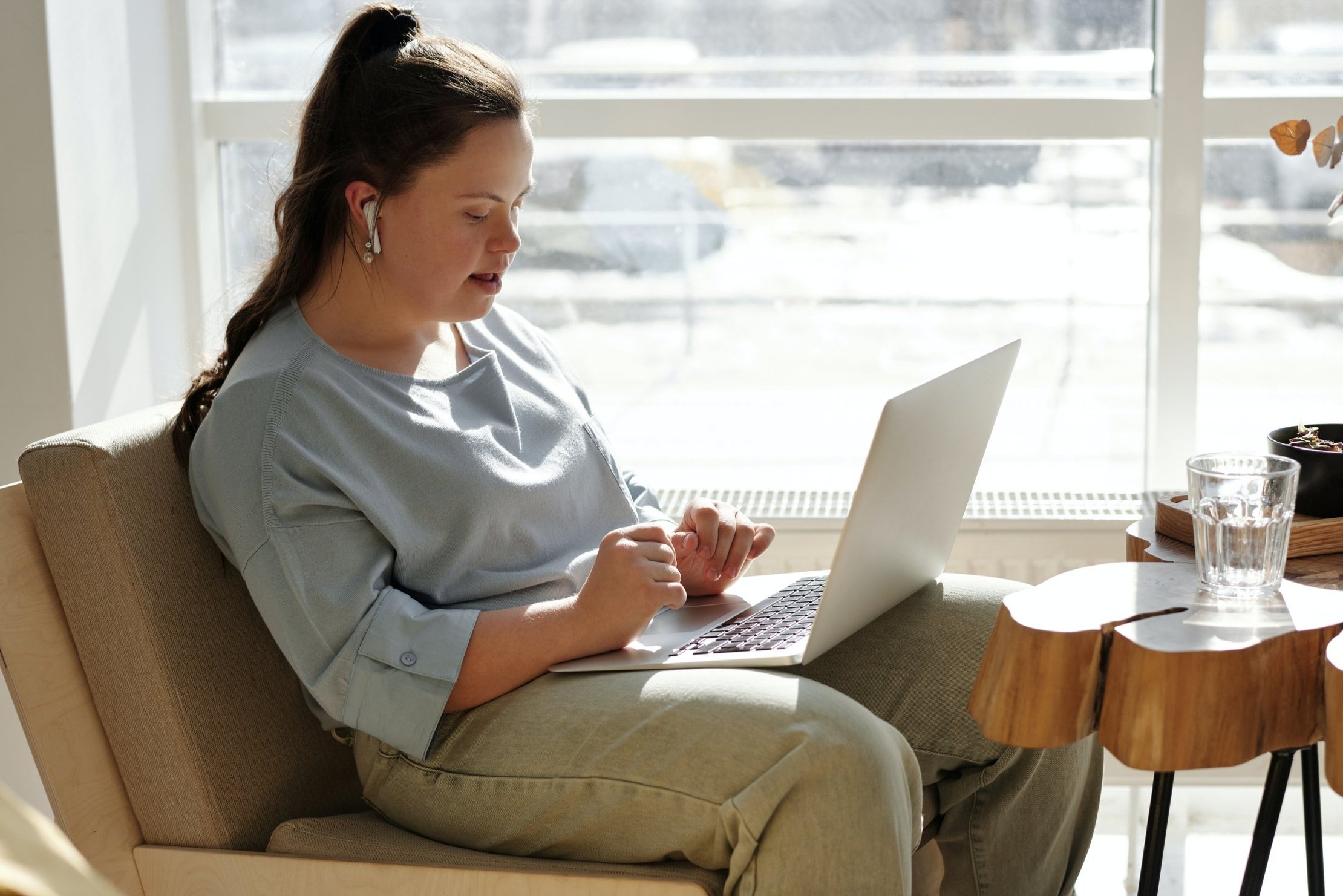 Person sitting on a chair with a laptop, wearing wireless earbuds, near a window and a wooden table with a glass of water.