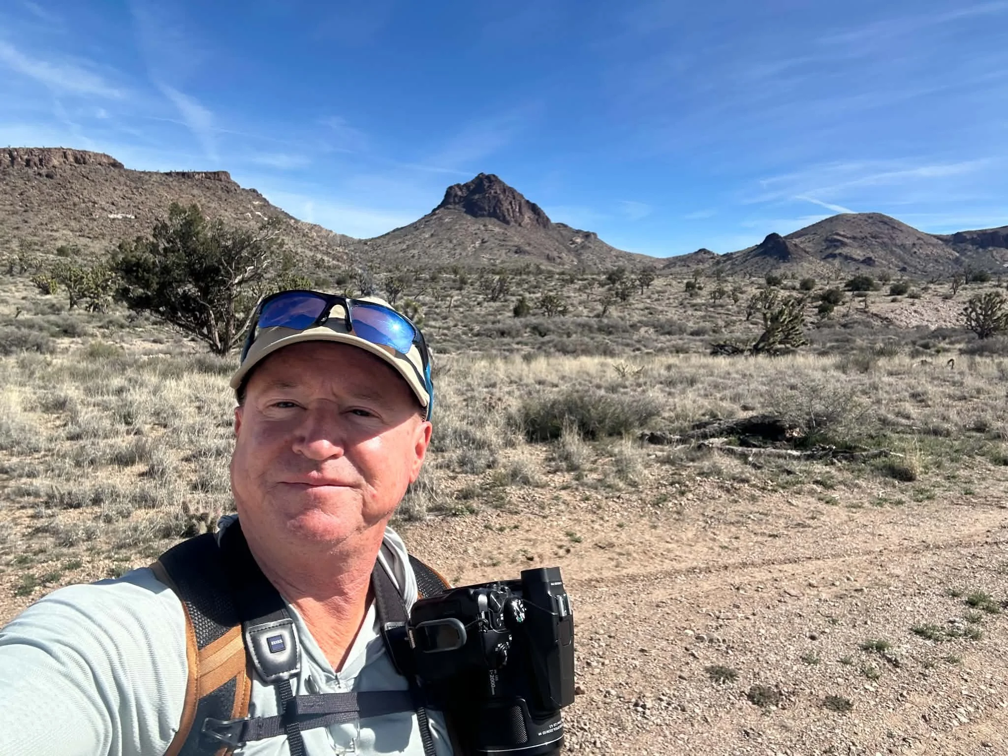A photo of the speaker, Tristen Davis, holding a camera. He is located in a remote desert area and wearing a hat with sunglasses.