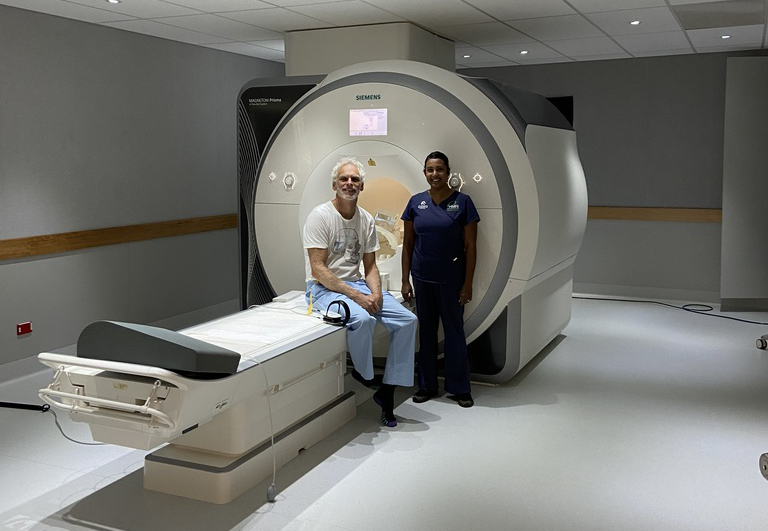A man sitting on the edge of a medical imaging machine with a nurse standing beside him in a hospital room.