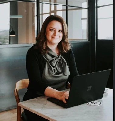 A woman sitting at a table with a laptop in a modern office or cafe setting, wearing a black cardigan and gray blouse with a bow, smiling at the camera.