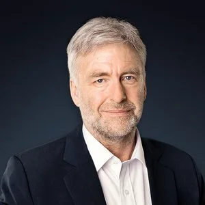 A senior man with gray hair and a beard, wearing a dark blazer and white shirt, posing against a dark backdrop.