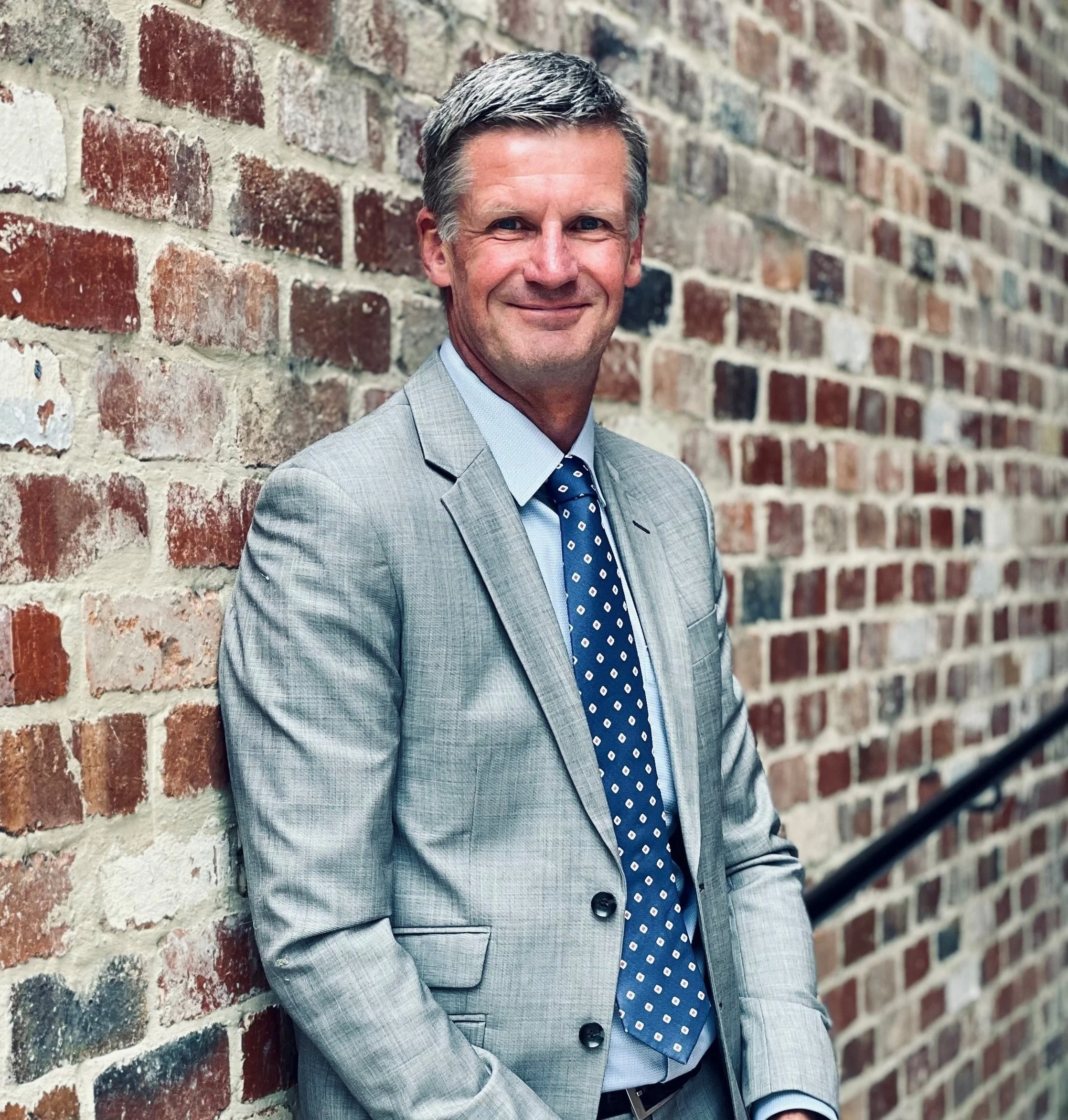 A professionally dressed man in a gray suit with a blue and white polka dot tie, smiling while leaning against a red brick wall.
