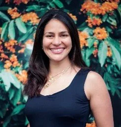 A woman smiling outdoors in front of orange flowers and green foliage, wearing a sleeveless dark blue top.