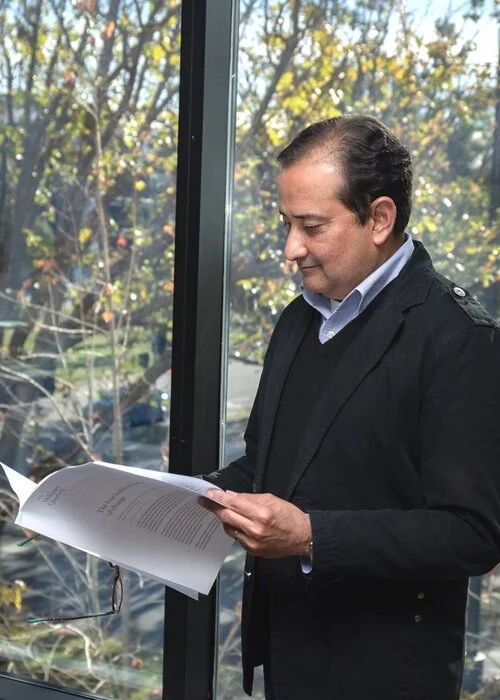 A man with dark hair, wearing a dark jacket and white shirt, is reading a document and standing by a window with trees outside.