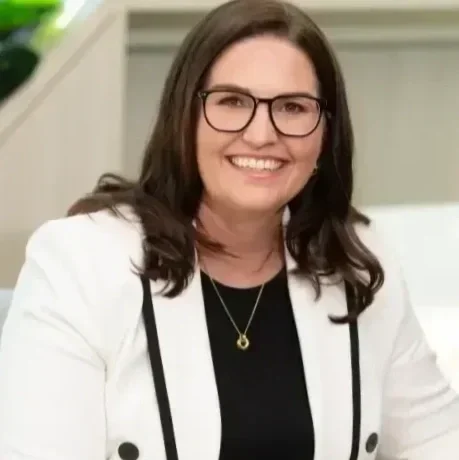 A woman with shoulder-length dark hair, glasses, and a white blazer smiling in an indoor setting.