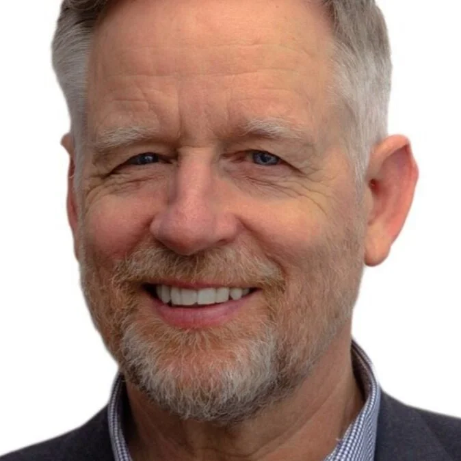 Close-up of a middle-aged man with short gray hair, a beard, and blue eyes, smiling and wearing a collared shirt and blazer.