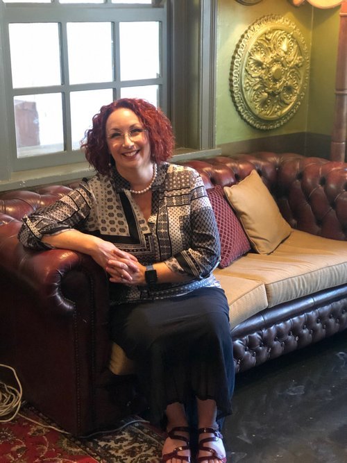 A woman with curly red hair sitting on a brown leather sofa in a cozy room, smiling at the camera. The room has a window with divided panes, a decorative wall piece, and colorful pillows on the sofa.