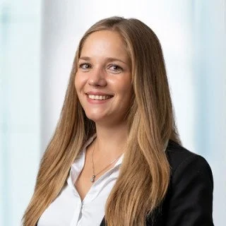 Portrait einer lachenden jungen Frau mit langen, glatten, hellbraunen Haaren, weißem Hemd und schwarzer Jacke, vor hellem Hintergrund.