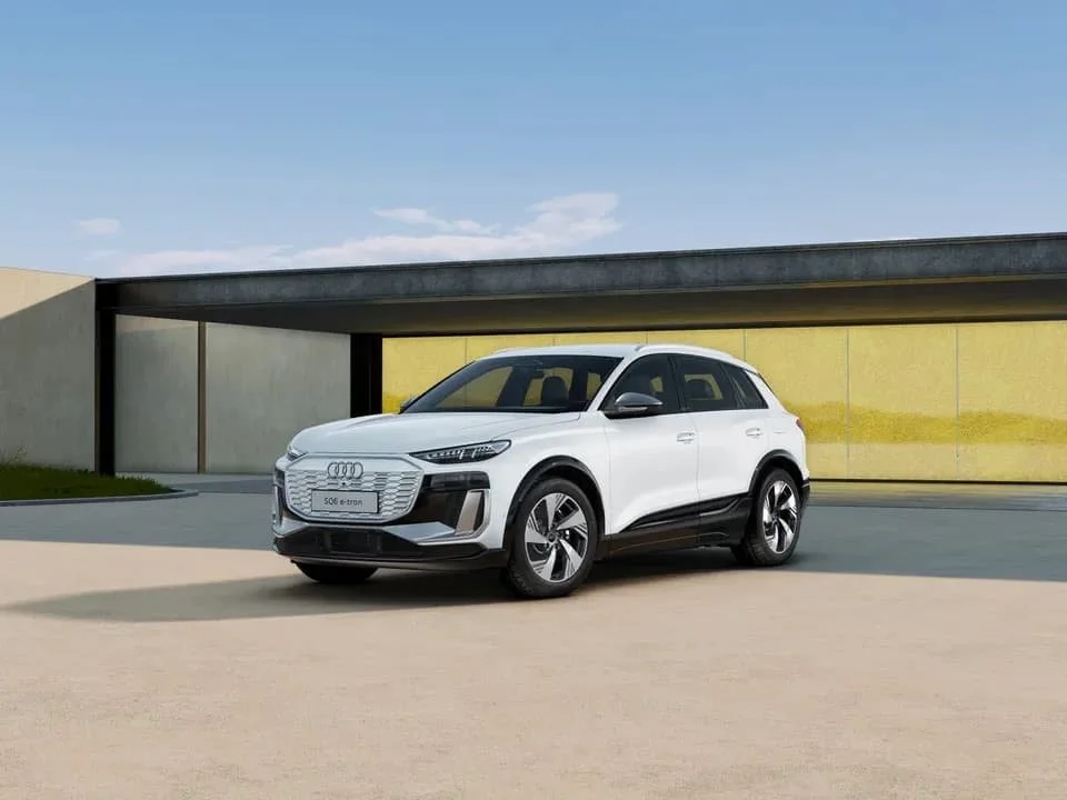 Ein weißer Audi Q4 e-tron Elektroauto vor einem modernen Gebäude mit gelber Wand und blauer Himmel.