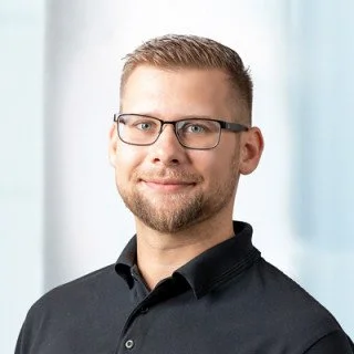 Porträt eines Mannes mit Brille und kurzer, heller Frisur, in dunklem Hemd, vor einem hellen Hintergrund.