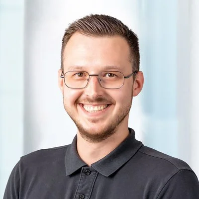 Porträt eines lachenden Mannes mit Brille, dunkles Polo-Shirt, vor einer weißen Vorhang-Hintergrund.