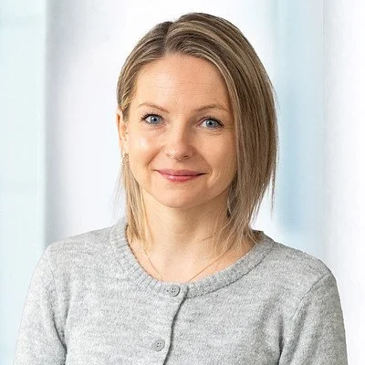 Porträt eines lachenden jungen Frau mit braunen Haaren, trägt eine grüne Bluse, vor einem weißen Hintergrund.