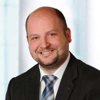 Porträt eines lächelnden Mannes in formeller Kleidung, mit dunklem Anzug und Krawatte, vor lichtblauem Hintergrund.