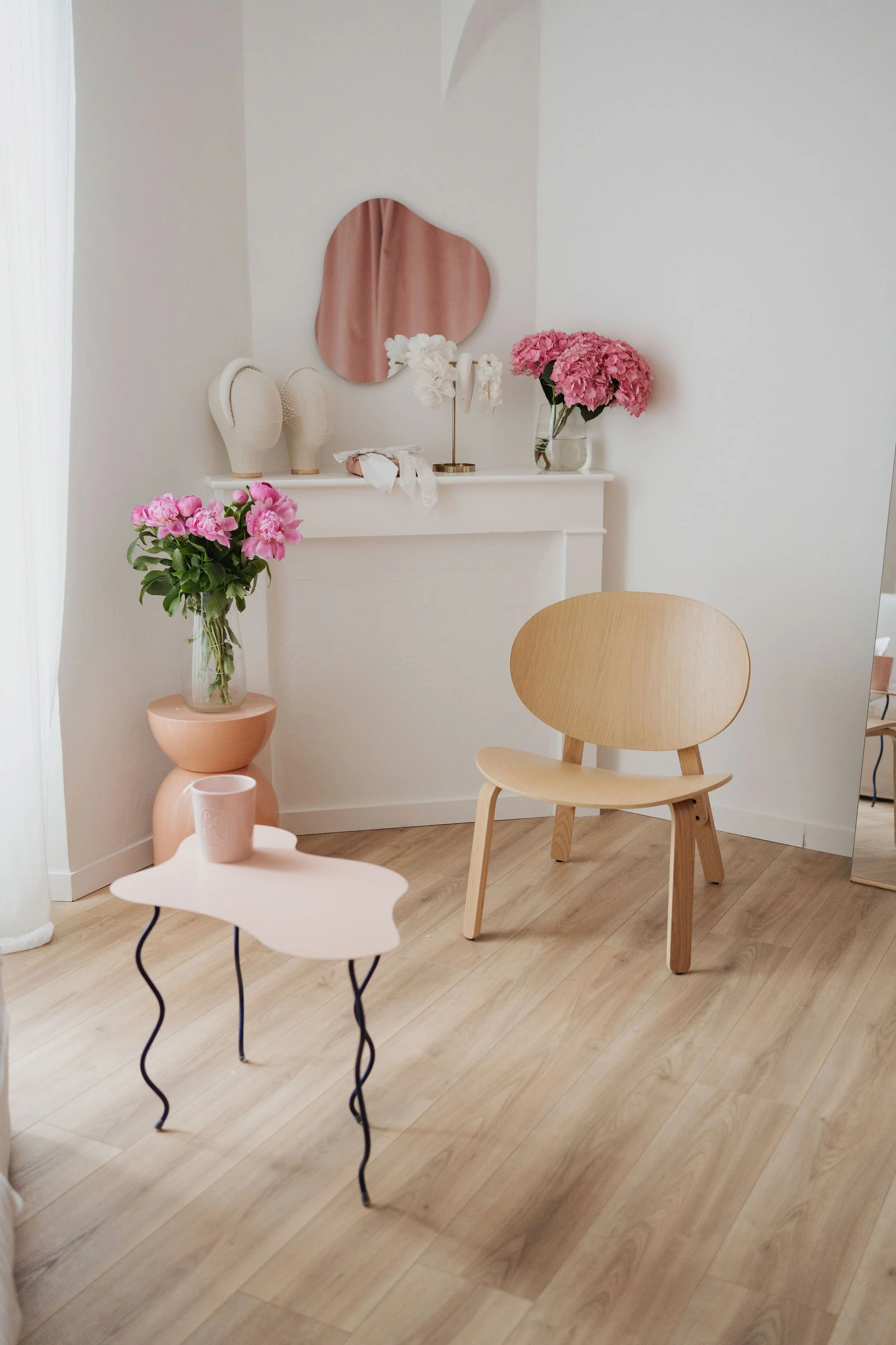Intérieur minimaliste avec une chaise en bois, une petite table rose, un vase avec des fleurs roses, un miroir, et des décorations sur une cheminée blanche.