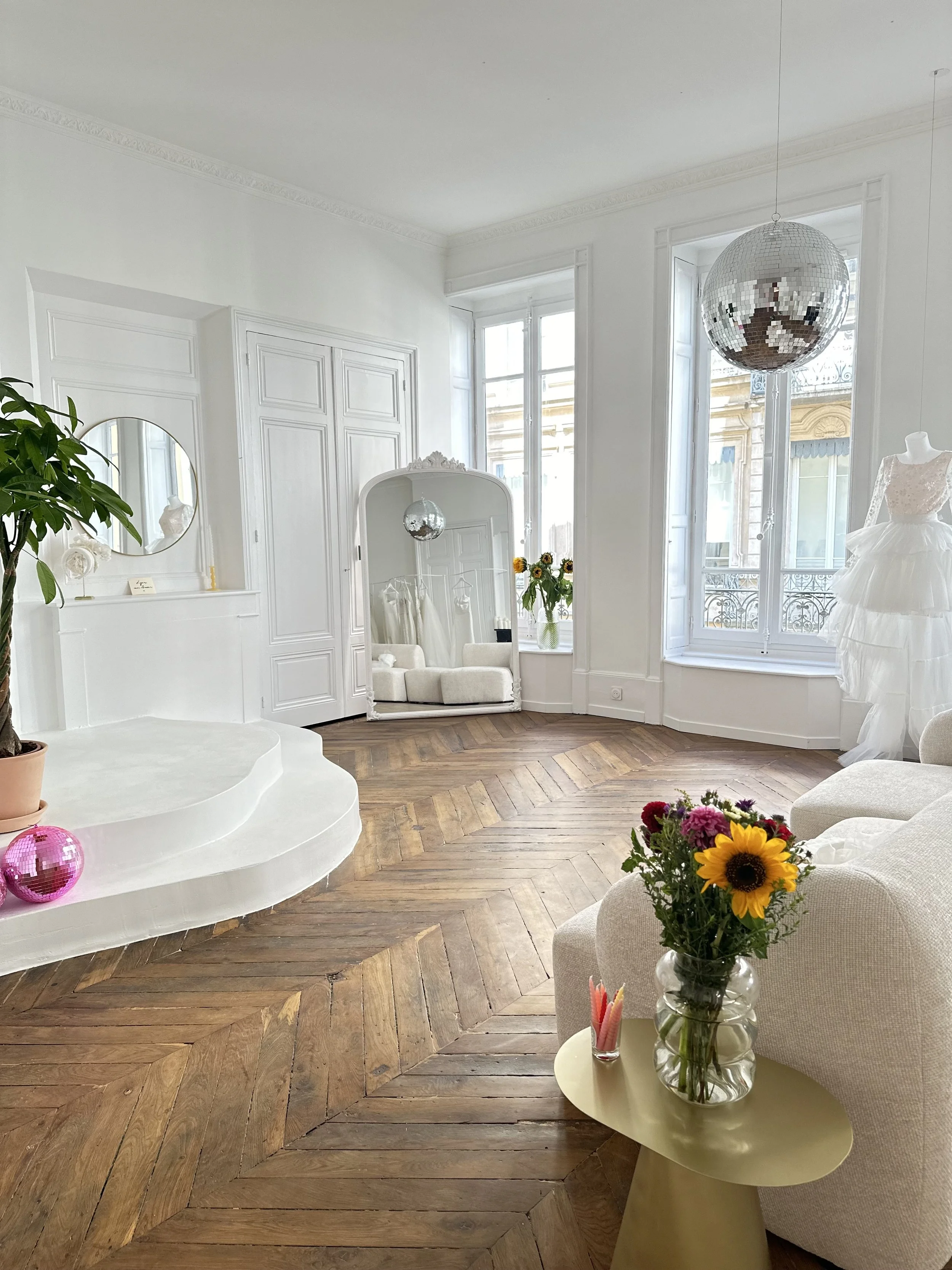 Salon lumineux avec parquet en bois, grand miroir, décoration blanche, chandelier disque miroir, fauteuils blancs, fleurs dans un vase, robe blanche sur mannequin.