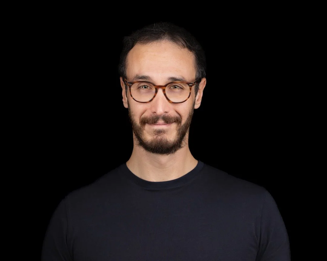 A man with glasses and a beard smiling against a black background.