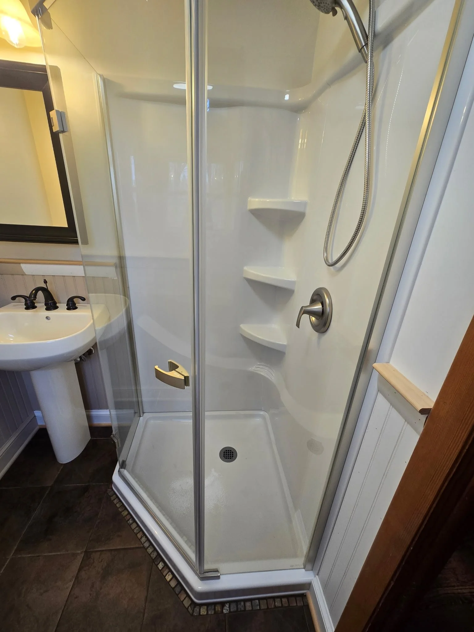 A small white shower stall with a sliding glass door, built-in shelves, a handheld showerhead, and a brushed nickel handle, next to a bathroom sink with a black faucet.