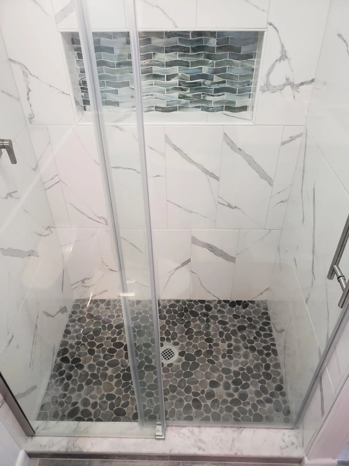 A small walk-in shower with white marble-like tiles on the walls, a built-in niche with gray and black mosaic tiles, a pebble stone floor, and a metal drain in the center.