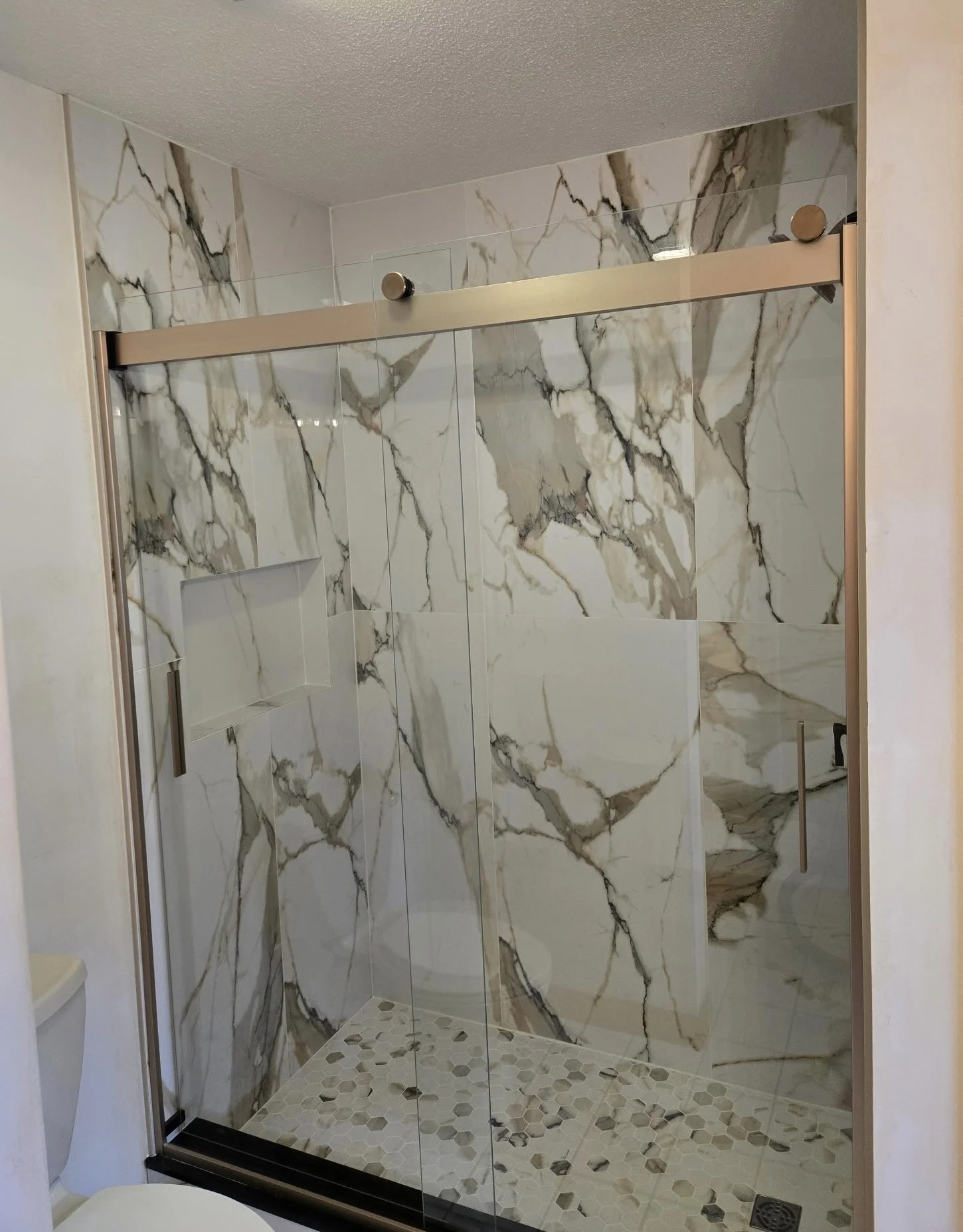 A modern walk-in shower with glass walls and marble tile walls with gray, black, and beige veining, and a hexagonal tile floor.