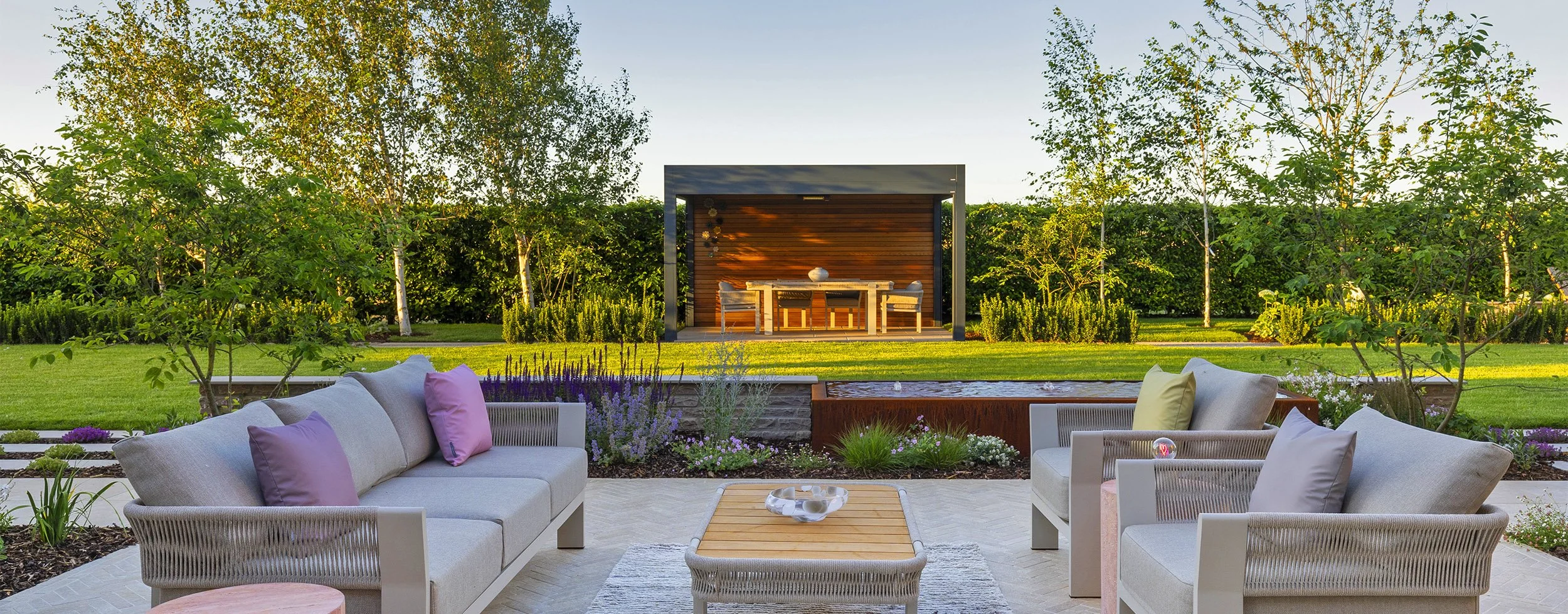 A garden patio with outdoor seating, herringbone paving, a wooden table in front overlooking a green lawn with trees and a small outdoor dining area with a table and chairs under a louvered Aluminium pergola.