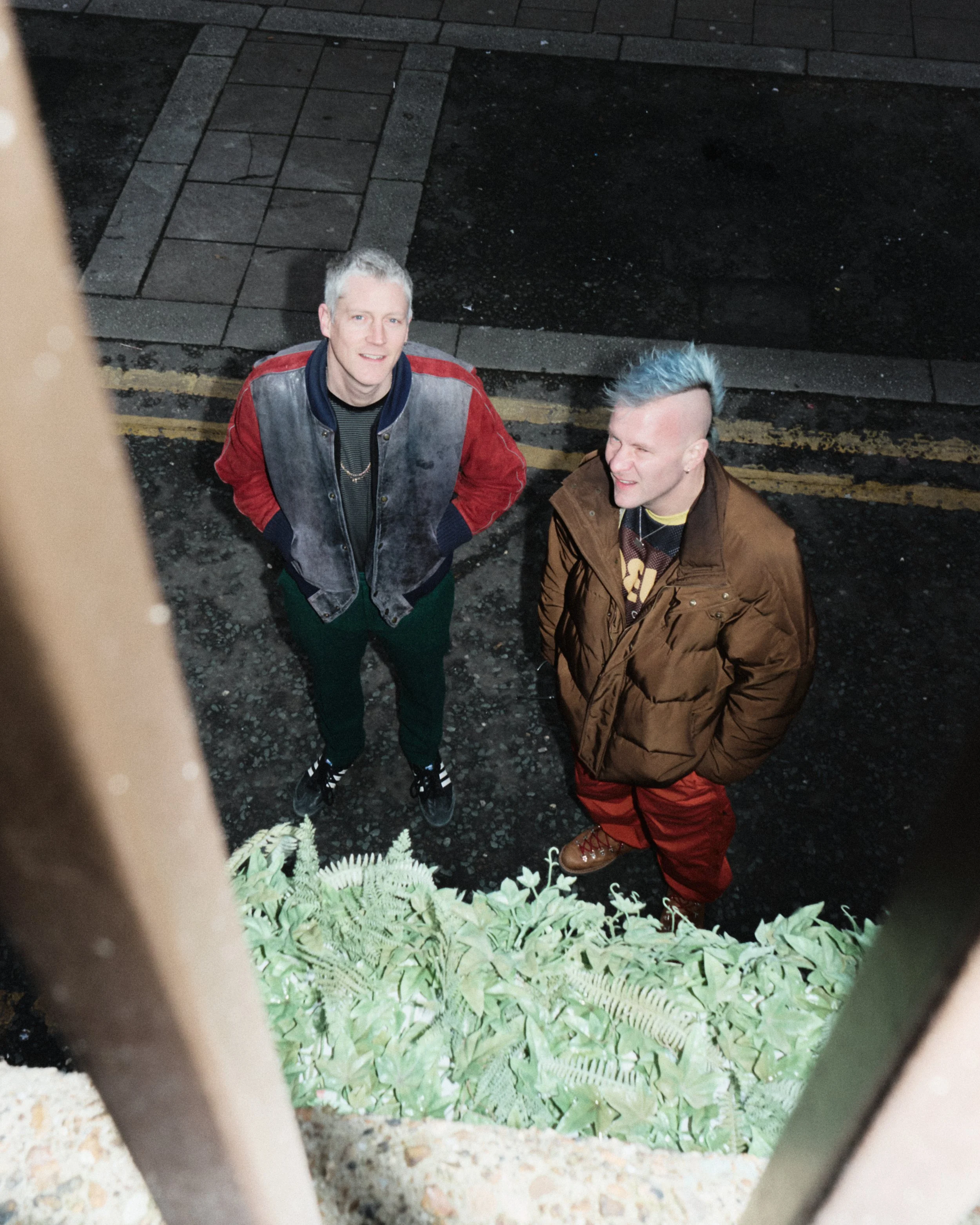 Two men standing outside on a dark street, looking up at the camera, with plants in the foreground.