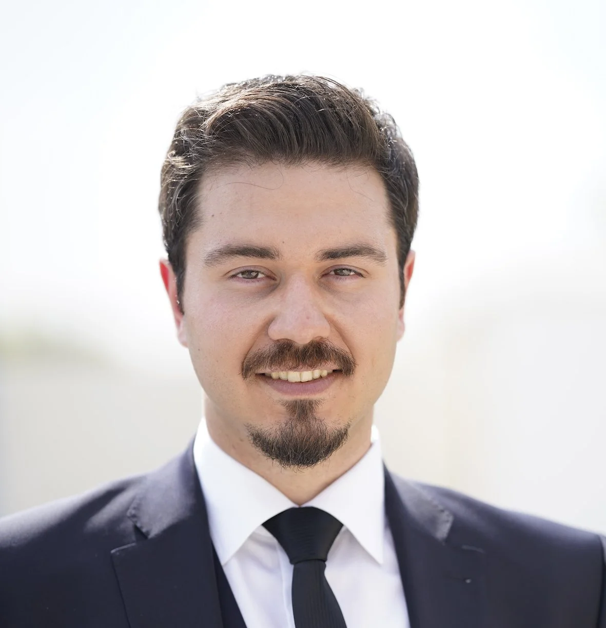 Portrait of a man with dark hair, mustache, and goatee, wearing a dark suit, white shirt, and black tie, smiling slightly against a bright, blurred outdoor background.