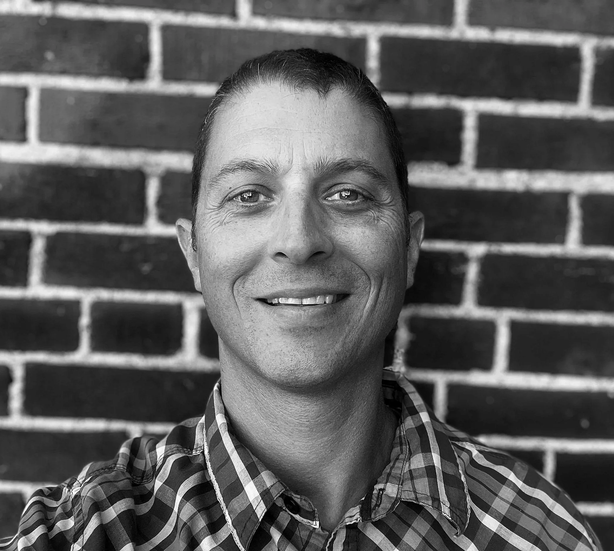 A smiling person with short hair and a checkered shirt, looking up, standing in front of a brick wall.