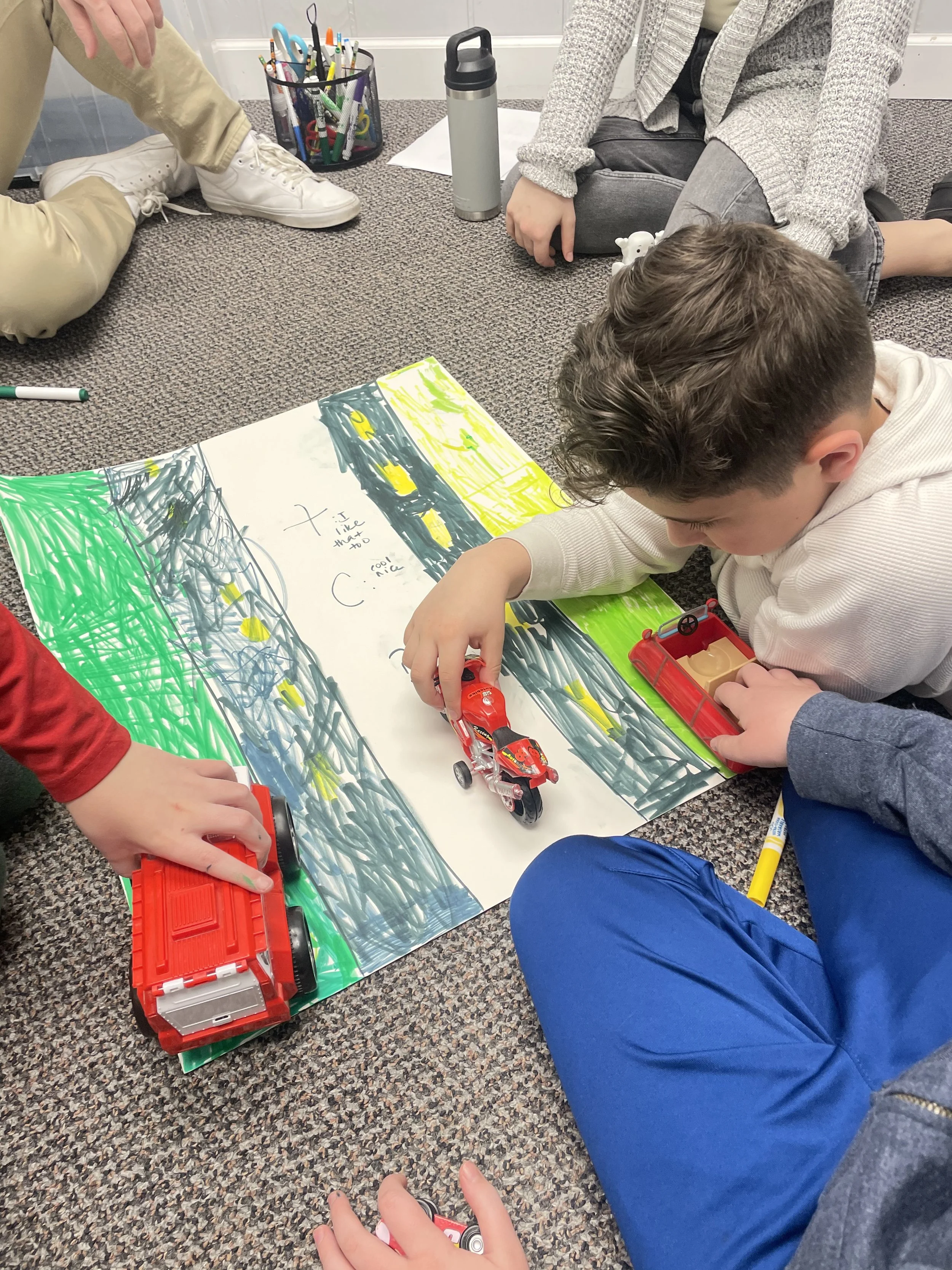 Children sitting on a carpeted floor playing with toy cars on a large piece of paper with colorful drawings.