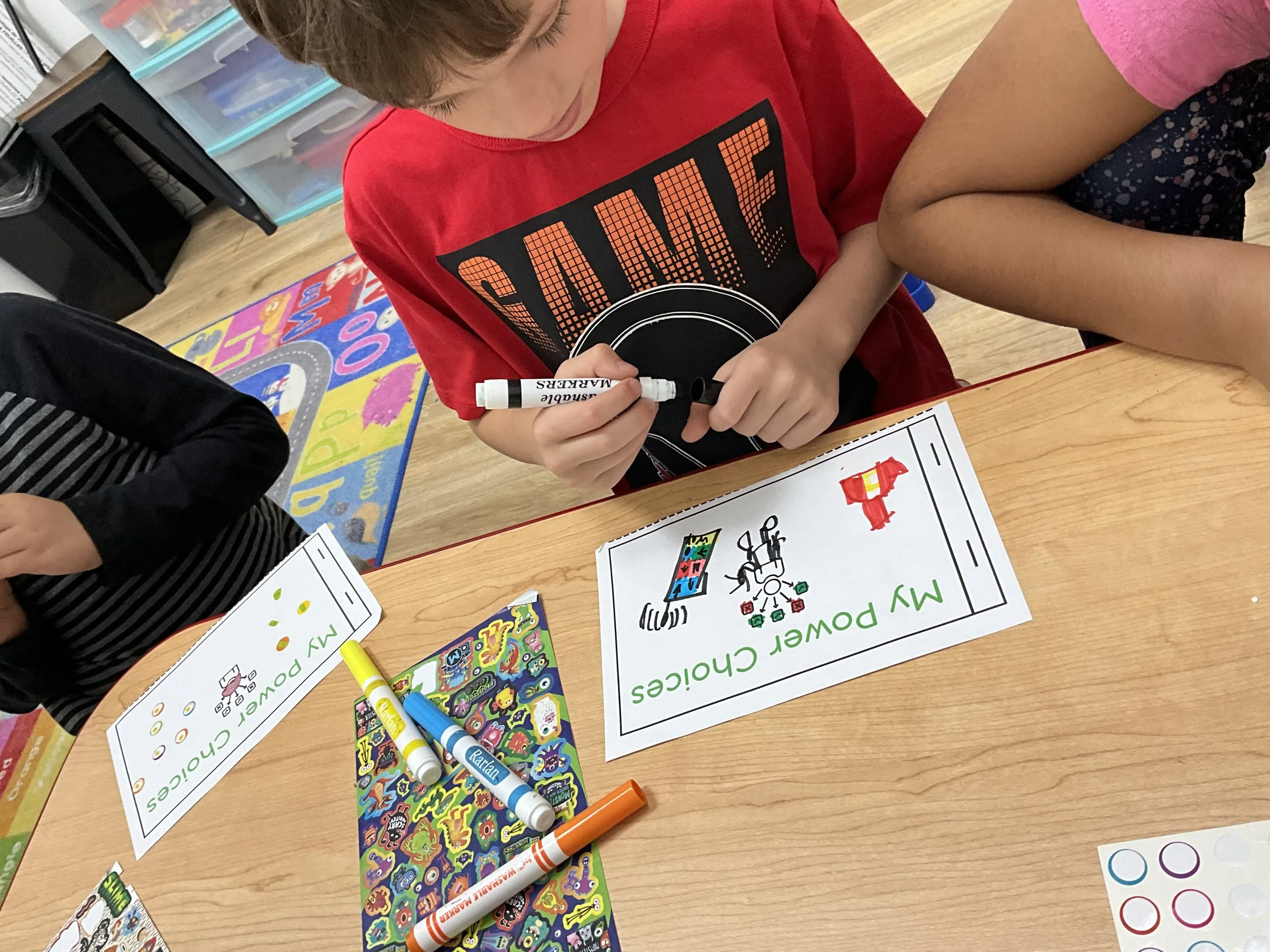 A child in a red shirt drawing on a worksheet with markers at a classroom table, with colorful stickers, markers, and learning materials around.