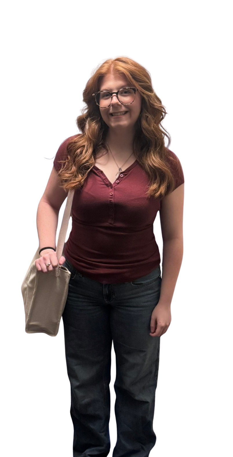 Young woman with red hair, glasses, wearing a maroon t-shirt and black jeans, carrying a beige shoulder bag, smiling.