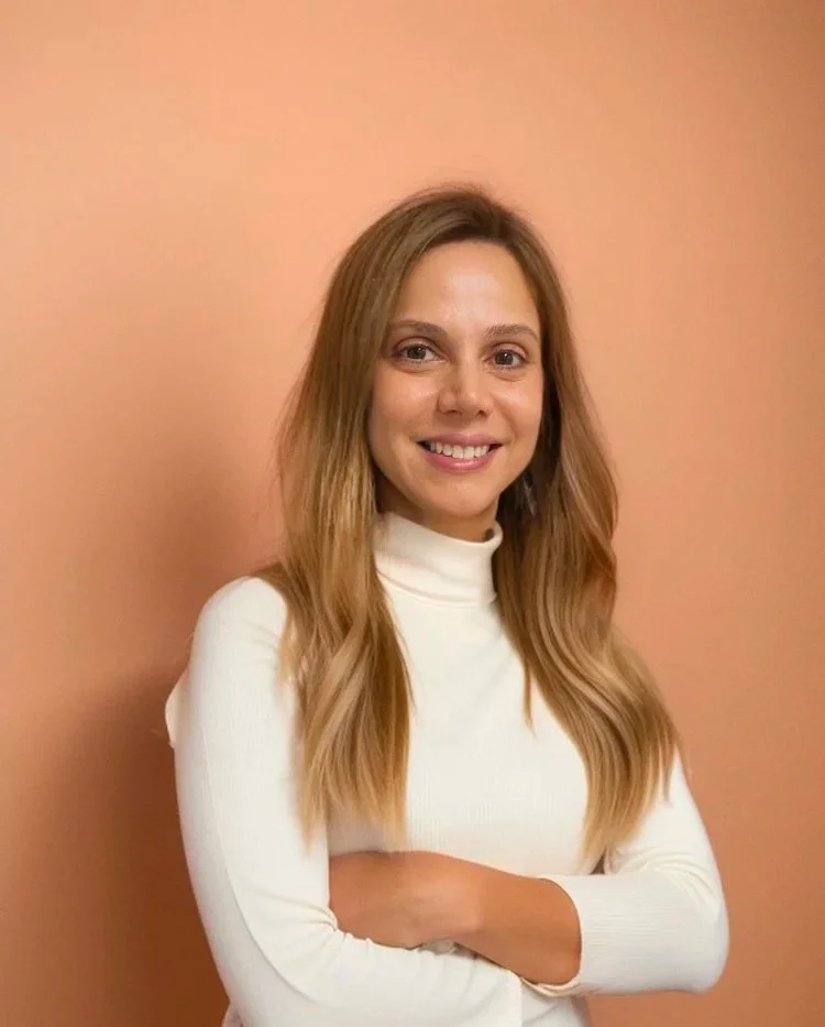A professional speech-language pathologist with long, wavy blonde hair wearing a white turtleneck sweater, smiling with her arms crossed, standing against a peach-colored wall.