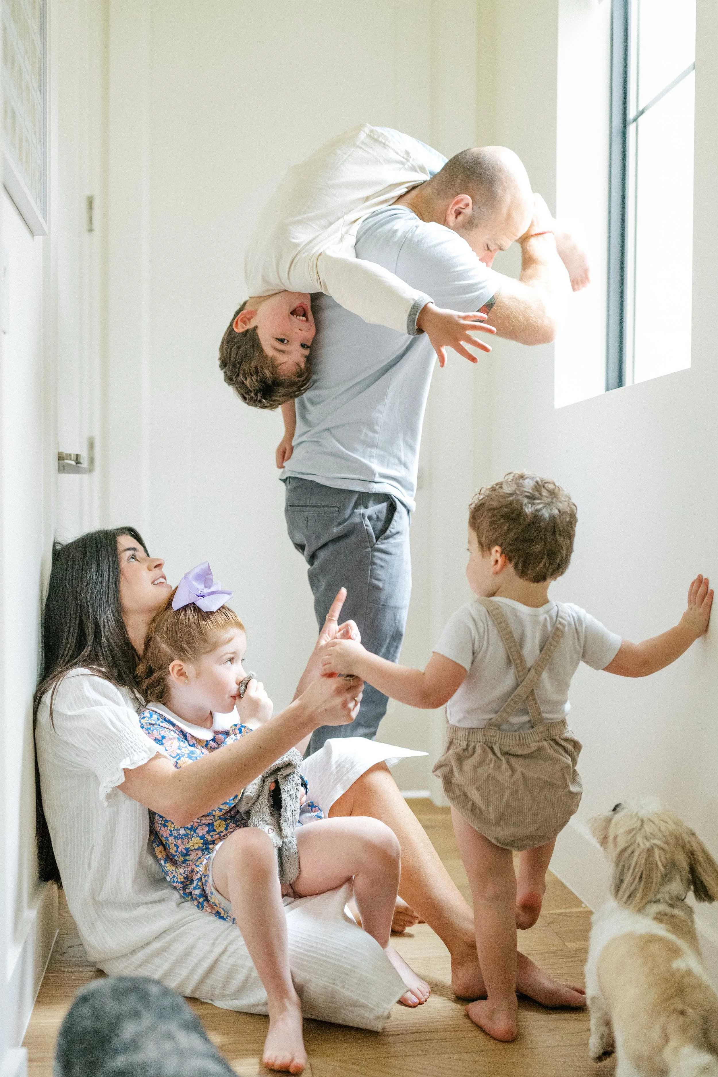 Mom sitting on the ground, cuddling two littles, and dad standing up holding a child upside down laughing during an in-home family session in Austin, Texas.