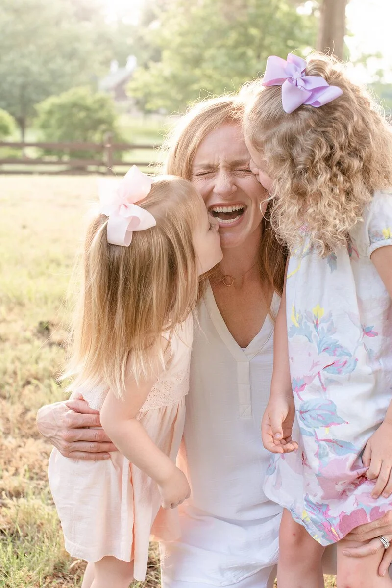 Mom kneeling on the ground during a beautiful summer evening with her daughters on both sides of her and her girls are kissing her checks while she laughs by Kelly Bullock Photography.